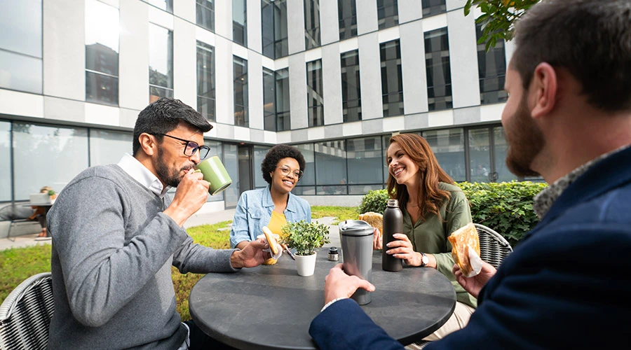 Gruppo di colleghi durante una pausa caffè in uno spazio esterno aziendale arredato, con tavolo e sedute in un cortile verde tra edifici per uffici.